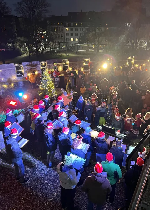 Foto vom Adventssingen auf der Burg: der Chor in Weihnachtsmützen vor einem beleuchteten Tannenbaum, dahinter das Publikum